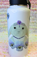 Cartoon gray lop-eared bunny, wearing fancy head jewelry and a rabbit constellation necklace, and surrounded by pansies, on a hydro-style tumbler.