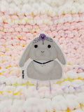 3-inch glossy vinyl sticker of a light grey Lop bunny, with a purple pansy and jewel head covering and a Lepus necklace.